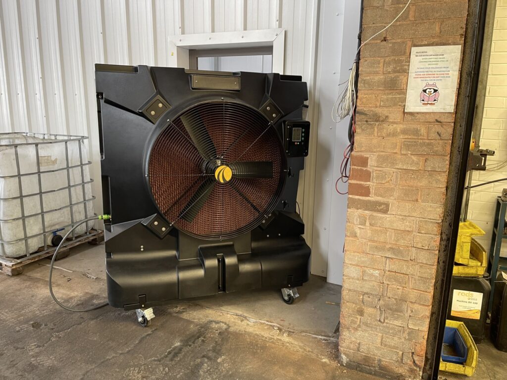 Large industrial fan on wheels next to a brick wall and a white container in a warehouse.