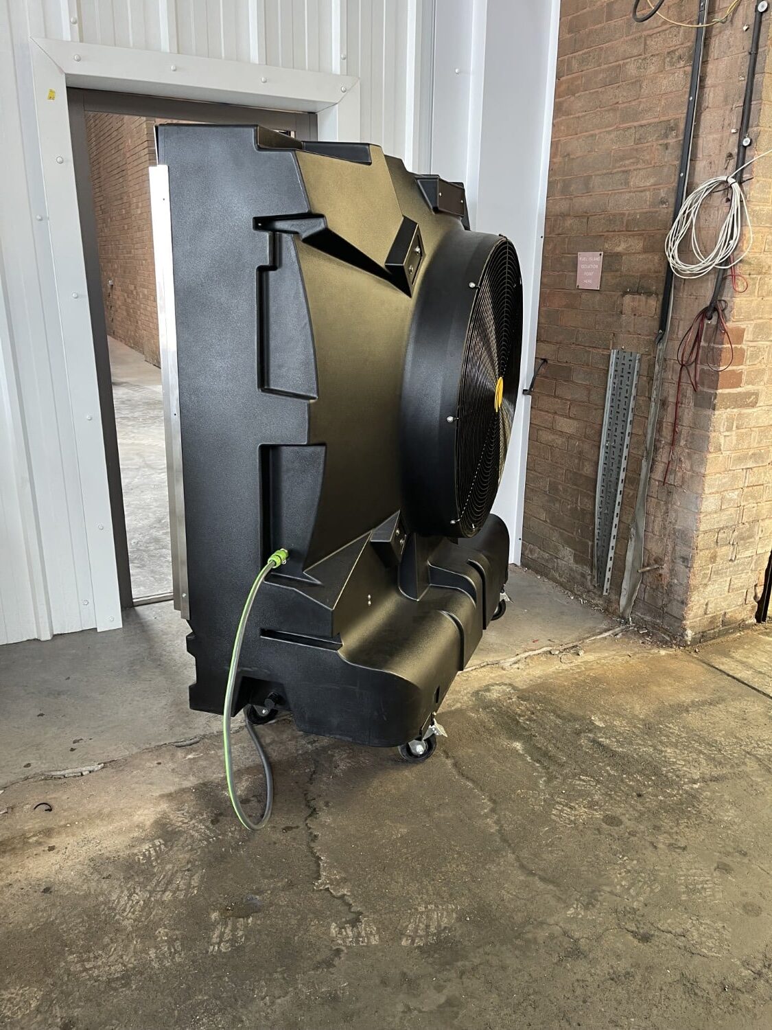 Large black industrial fan on wheels, plugged in, inside a warehouse near a brick wall and cables.