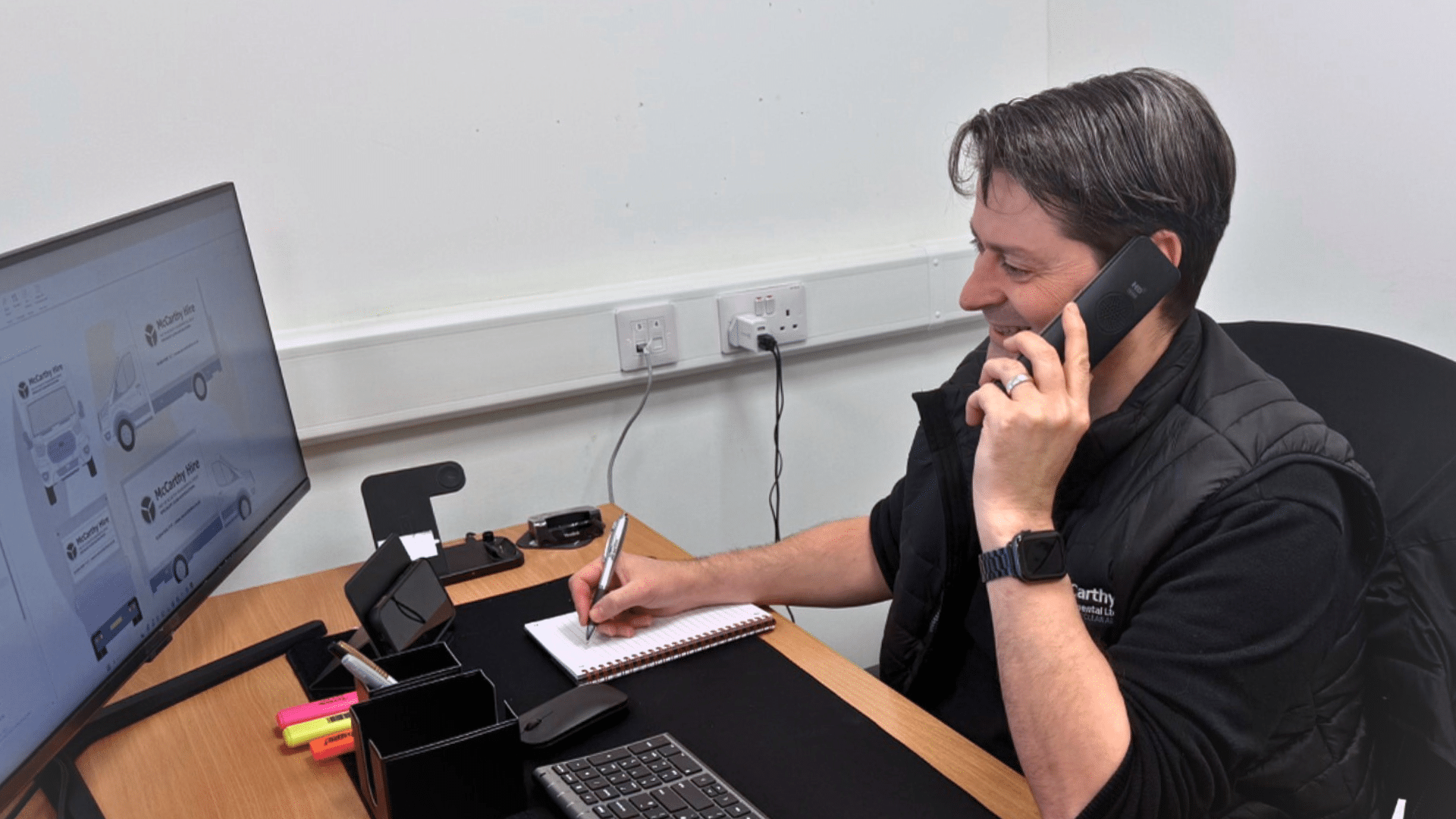 Man at a desk talking on the phone and writing notes, with a computer screen and office supplies.