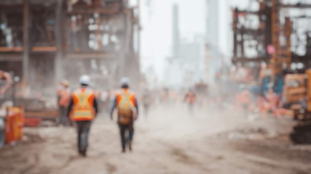 Workers in safety vests walk through a dusty construction site.