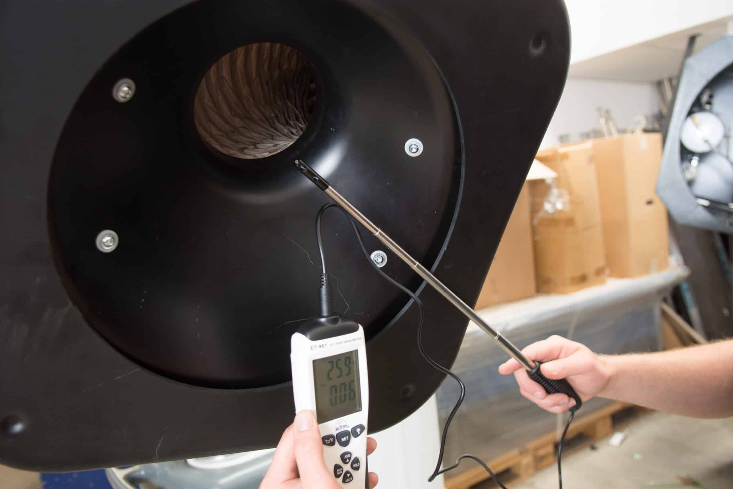 A person measures airflow with a digital anemometer inside a large industrial vent.